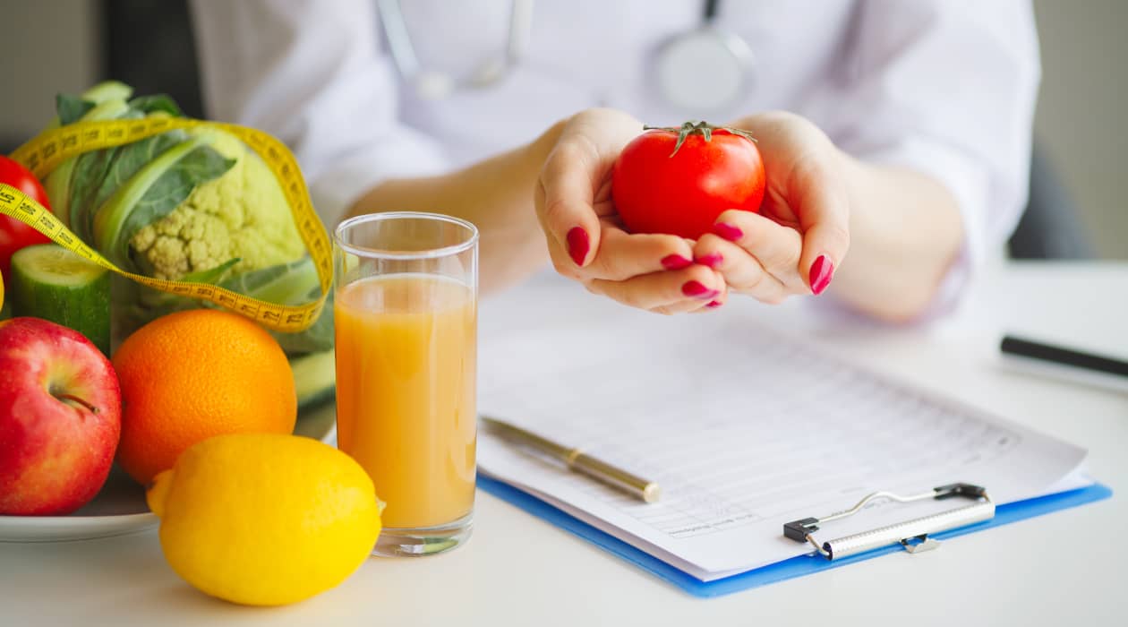 Nutricionista cogiendo un tomate entre sus manos y con frutas encima del escritorio