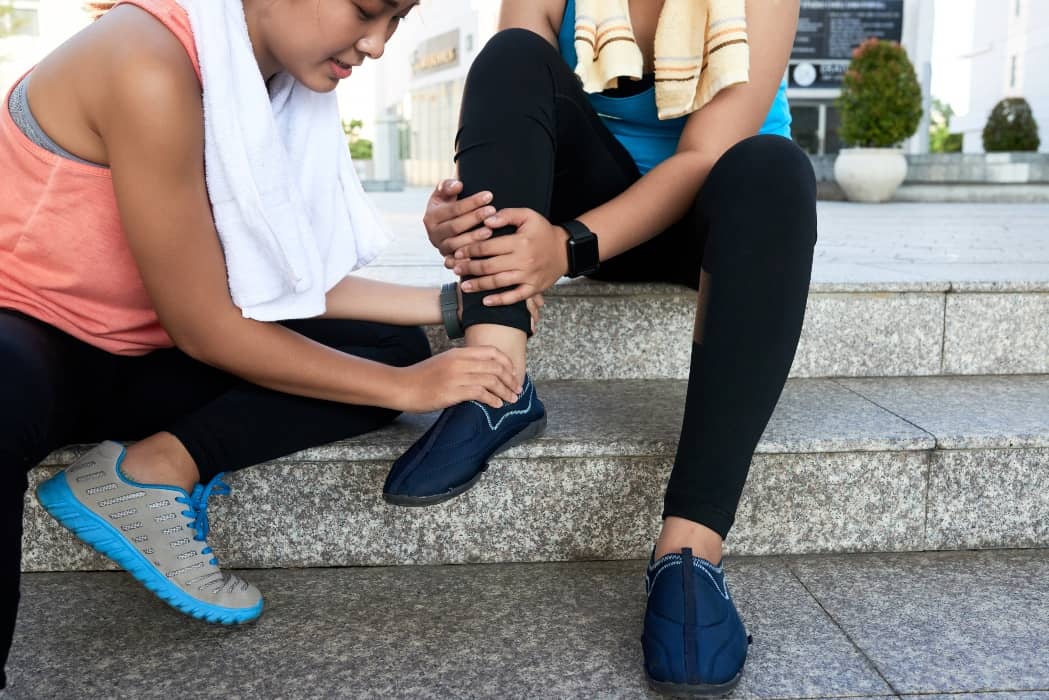 Mujer deportista apretando el tobillo de su compañera sentadas en unos escalones de la calle