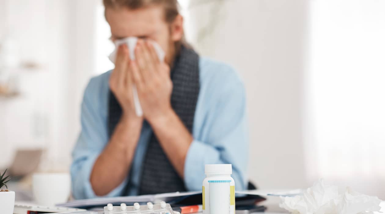 Primer plano de unos antibióticos y detrás un hombre estornudando limpiándose la nariz