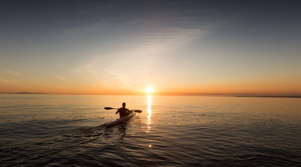 Hombre de espalda con su kayak en el mar mirando el atardecer