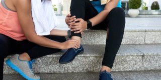 Mujer deportista apretando el tobillo de su compañera sentadas en unos escalones de la calle