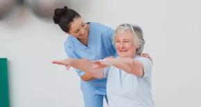 anciana sentada en un fitball con los brazos extendidos guiada por un técnico