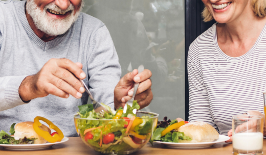 Hombre mayor sonriente moviendo una ensalada encima de una mesa al lado de una mujer