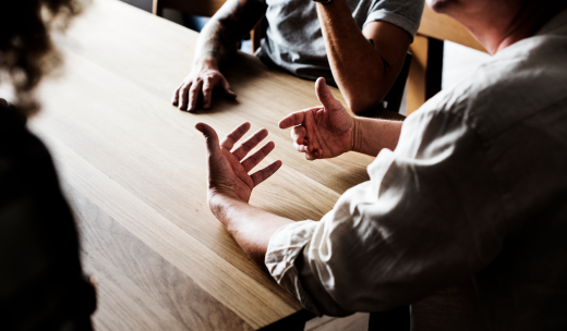 Tres personas sentadas en torno a una mesa gesticulando con las manos mientras discuten un tema