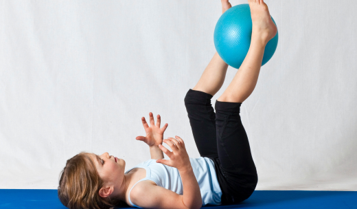Una niña pequeña tumbada en una esterilla con las piernas estiradas cogiendo una pelota