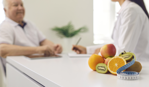 Primer plano de variedad de frutas y cinta métrica y detrás una especialista charlando con un hombre mayor
