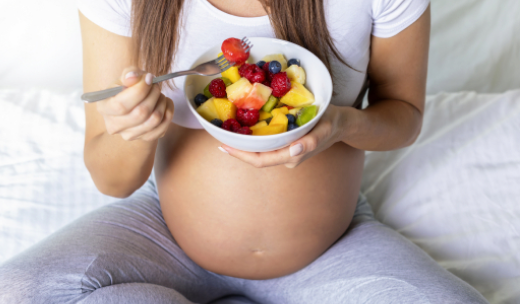 Una embarazada sentada con las piernas cruzadas comiendo un bol de frutas