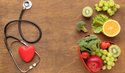 Mesa con frutas y verduras en un frutero con forma de corazón junto a un estetoscopio y un corazón de gomaespuma