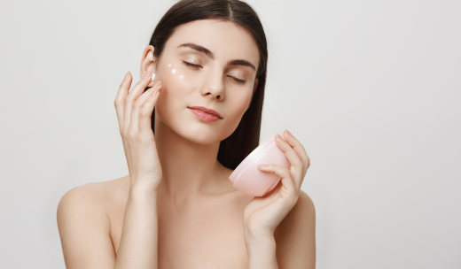 Mujer con los ojos cerrados echándose crema en la cara, sujetando el bote en una mano