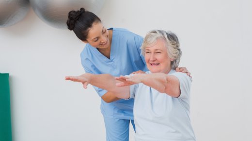 anciana sentada en un fitball con los brazos extendidos guiada por un técnico