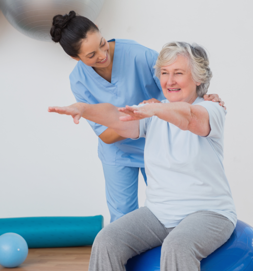 Anciana sentada sobre un fitball con los brazos extendidos con una profesional corrigiendo los ejercicios