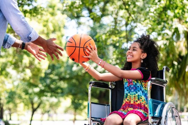 Niña sonriente en silla de ruedas pasándole una pelota de baloncesto a un adulto
