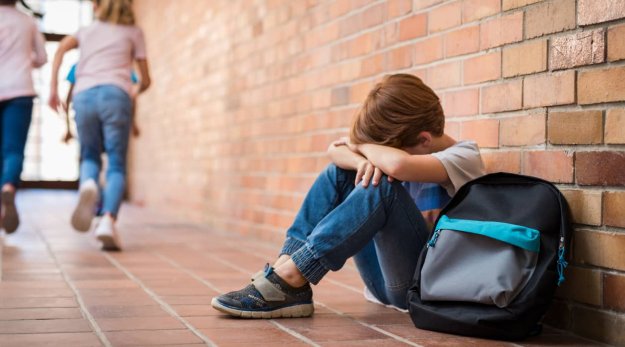 Niño sentado en el suelo del patio del colegio con la cabeza apoyada en las manos y una mochila al lado
