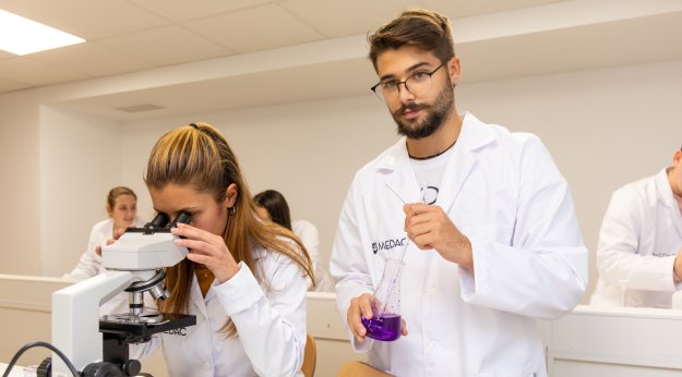 Alumnos con bata analizando muestras en un microscopio y una probeta